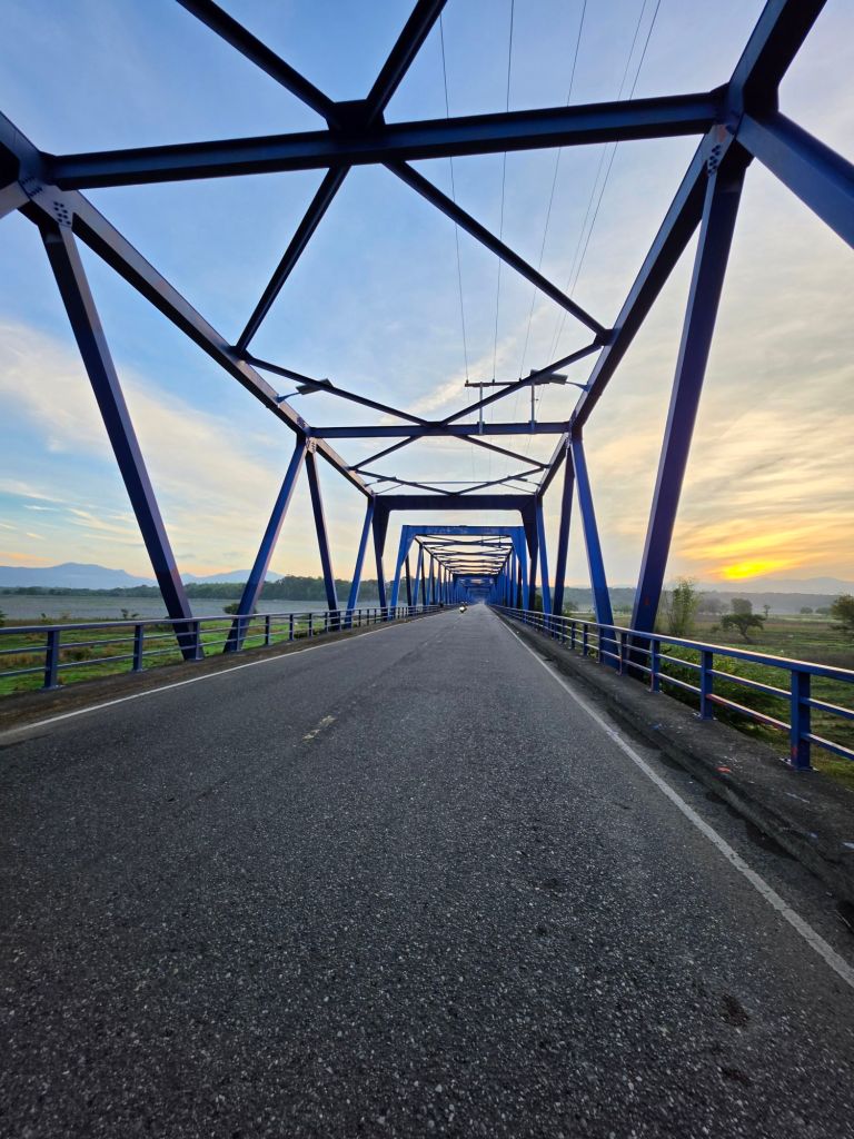 View of a blue truss bridge at sunrise, featuring a road running through the middle with lush greenery on either side.