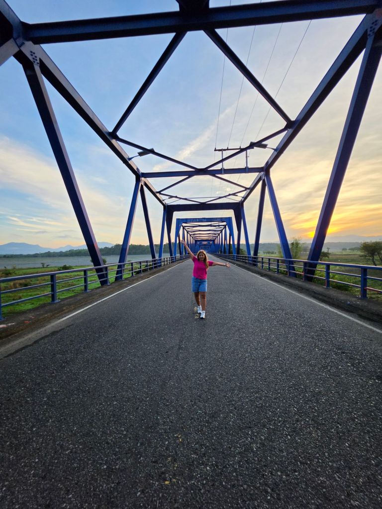 A person stands on an empty road bridging two sides, surrounded by a blue metal framework with a scenic background, during sunrise.