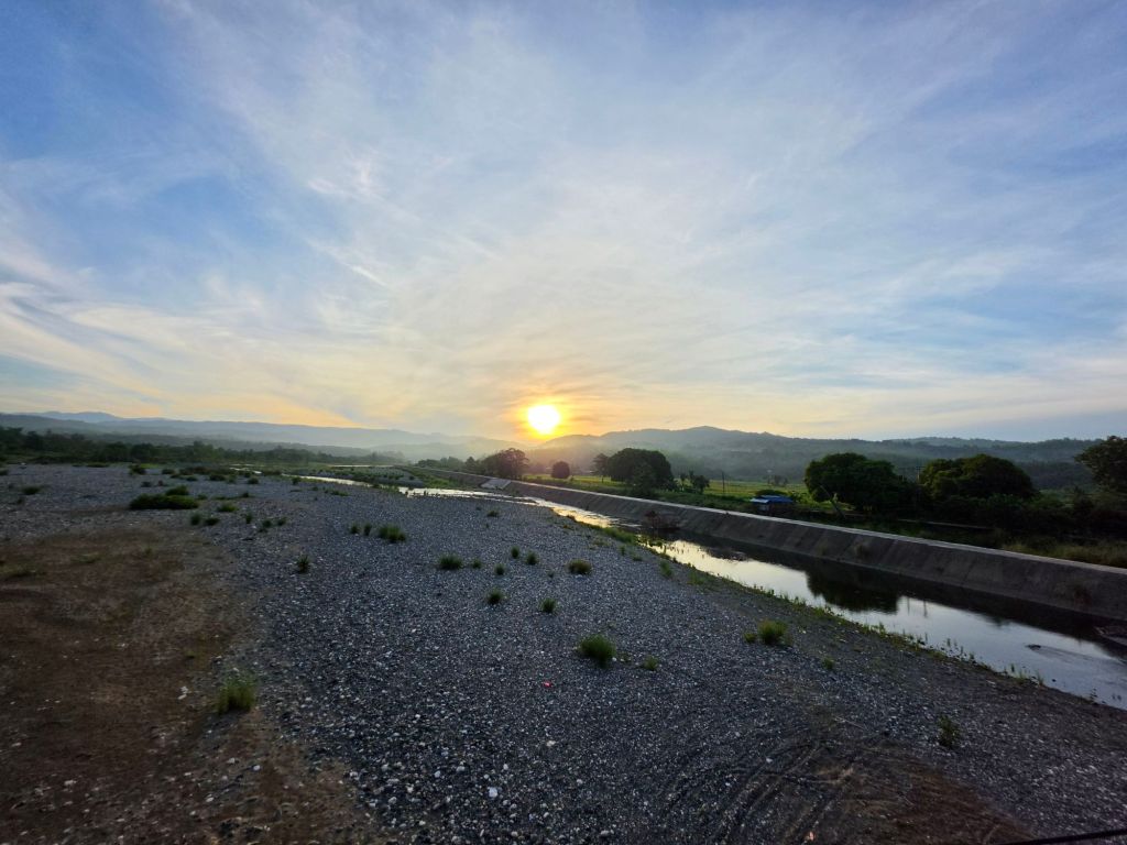 Sunset over a riverbank with rocky terrain and greenery in the background.