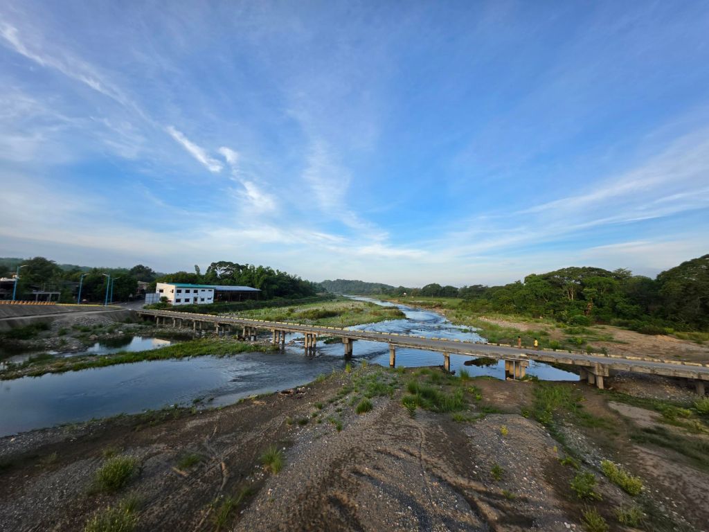 A tranquil river scene with a wooden bridge crossing over it, surrounded by lush greenery and trees under a blue sky with wispy clouds.