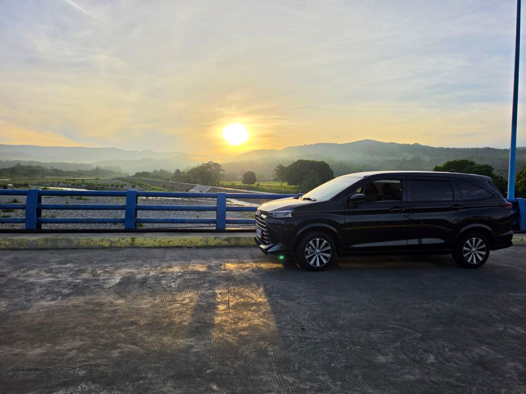 A black SUV parked on a road with a blue railing, set against a backdrop of a sunrise over mountains and a misty landscape.