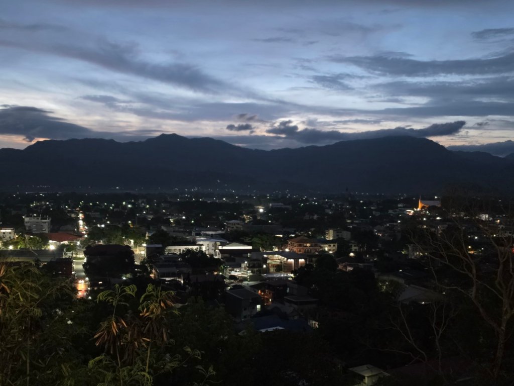 A nighttime cityscape view with illuminated buildings and streets, surrounded by mountains and a cloudy sky.