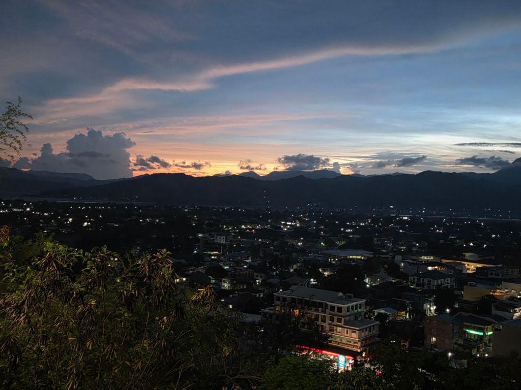 A panoramic view of a city at dusk, showcasing illuminated buildings and a colorful sunset with clouds and mountains in the background.