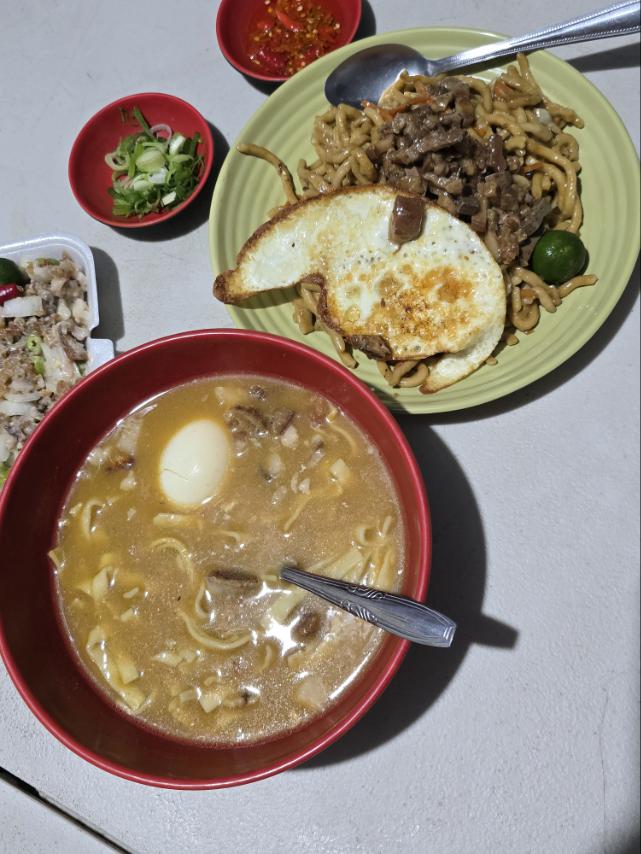 A bowl of steaming noodles in broth with a soft-boiled egg, accompanied by a plate of stir-fried noodles topped with beef and a fried egg, garnished with lime and green onions, with small bowls of red pepper sauce and chopped scallions on the side.