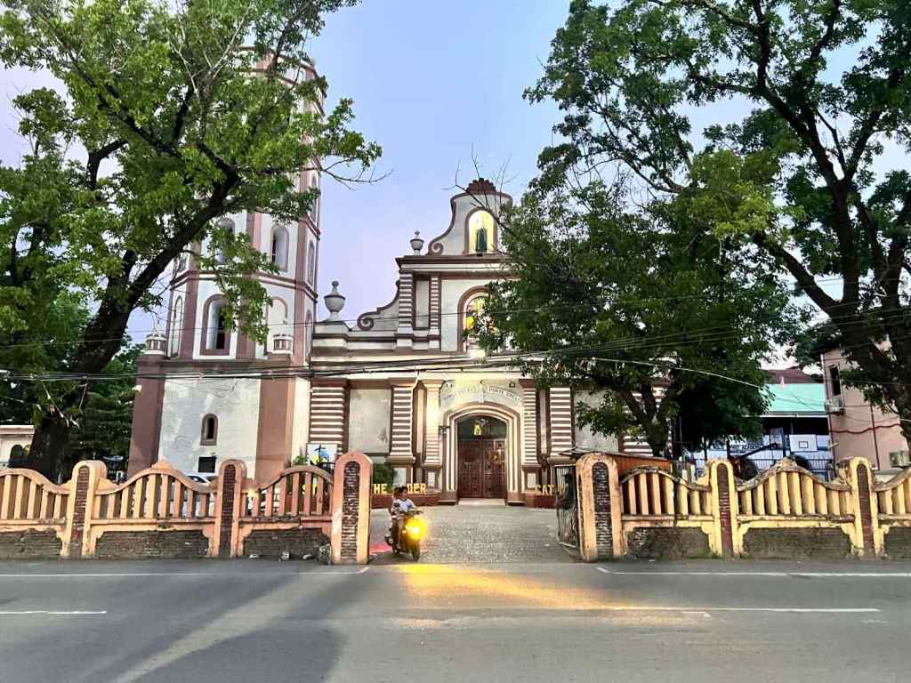 Image of a church with a unique architectural style, framed by large trees and a decorative fence, viewed from a road at dusk.