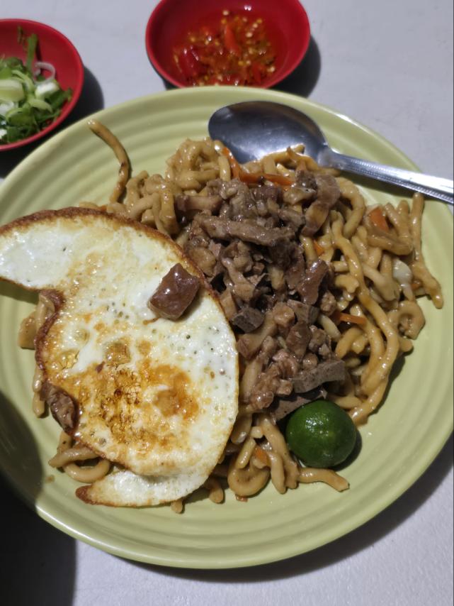 A plate of stir-fried noodles topped with diced beef and a sunny-side-up egg, accompanied by a lime and small bowls of sliced green onions and spicy salsa.